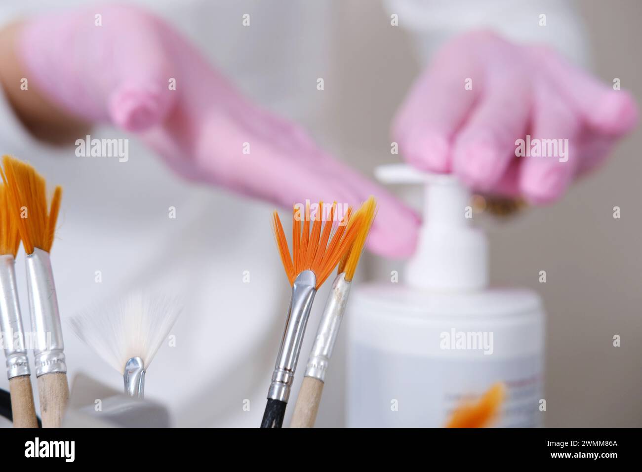 Eine Auswahl von Kosmetikbürsten im Fokus, wobei Hände Kosmetikgel für Gesichtsbehandlungen im Hintergrund abgeben. Stockfoto