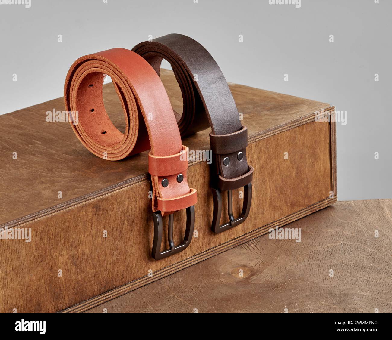 Zwei gefaltete, stilvolle Ledergürtel mit DAD-Prägung und matten Metallschnallen auf Holztreppen. Modisches personalisiertes Accessoire für Herren Stockfoto