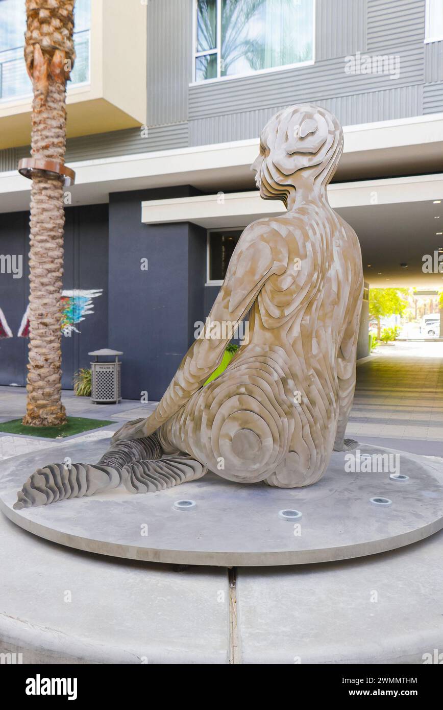 Skulptur einer Meerjungfrau, die auf einem runden Podest sitzt, in einem urbanen Außenbereich mit moderner Architektur im Hintergrund Stockfoto