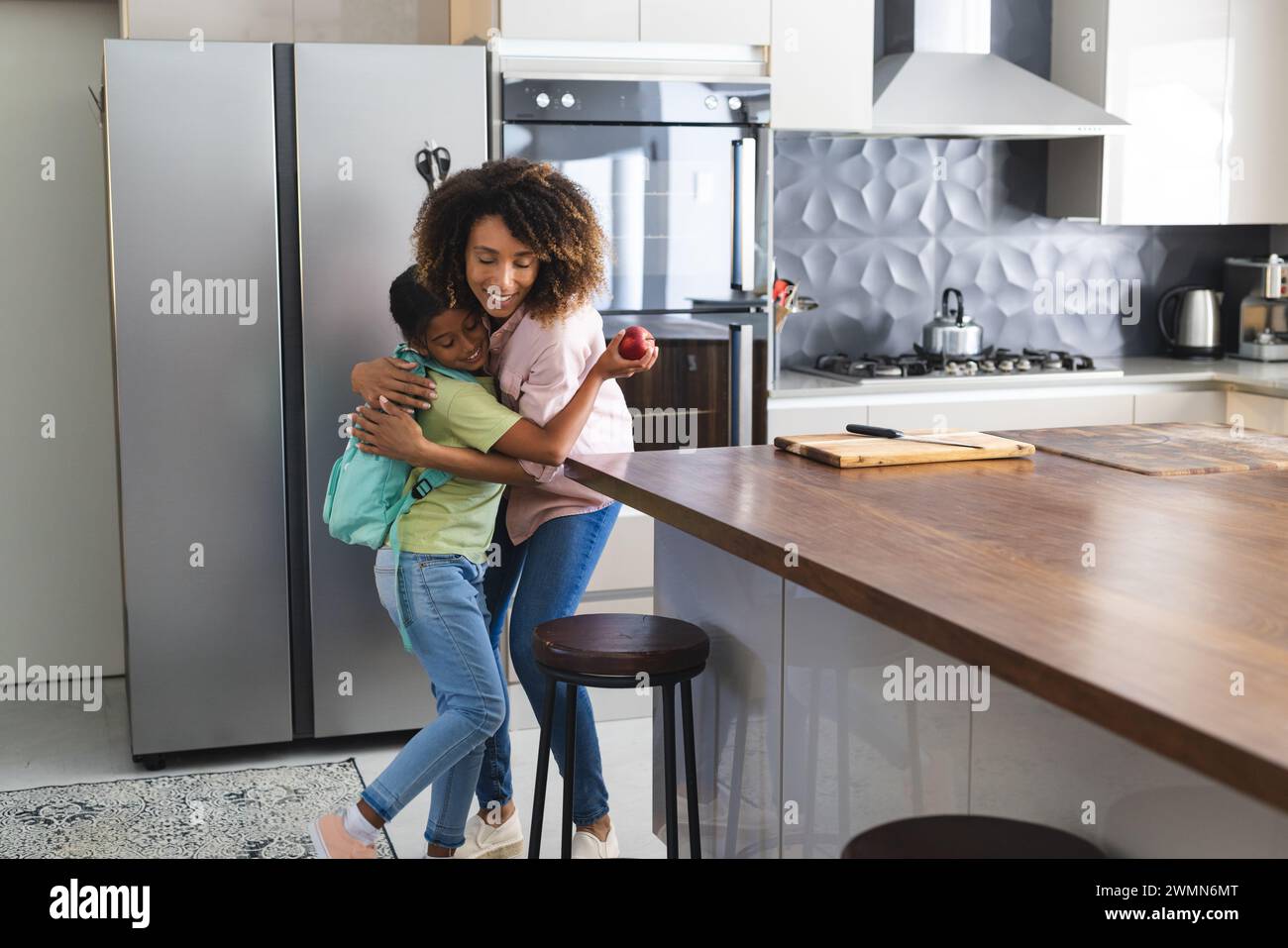 Birassische Mutter umarmt ihre birassische Tochter in einer modernen Küche. Sie teilen sich eine warme Umarmung und betonen eine pflegende Umgebung für die Tochter. Stockfoto