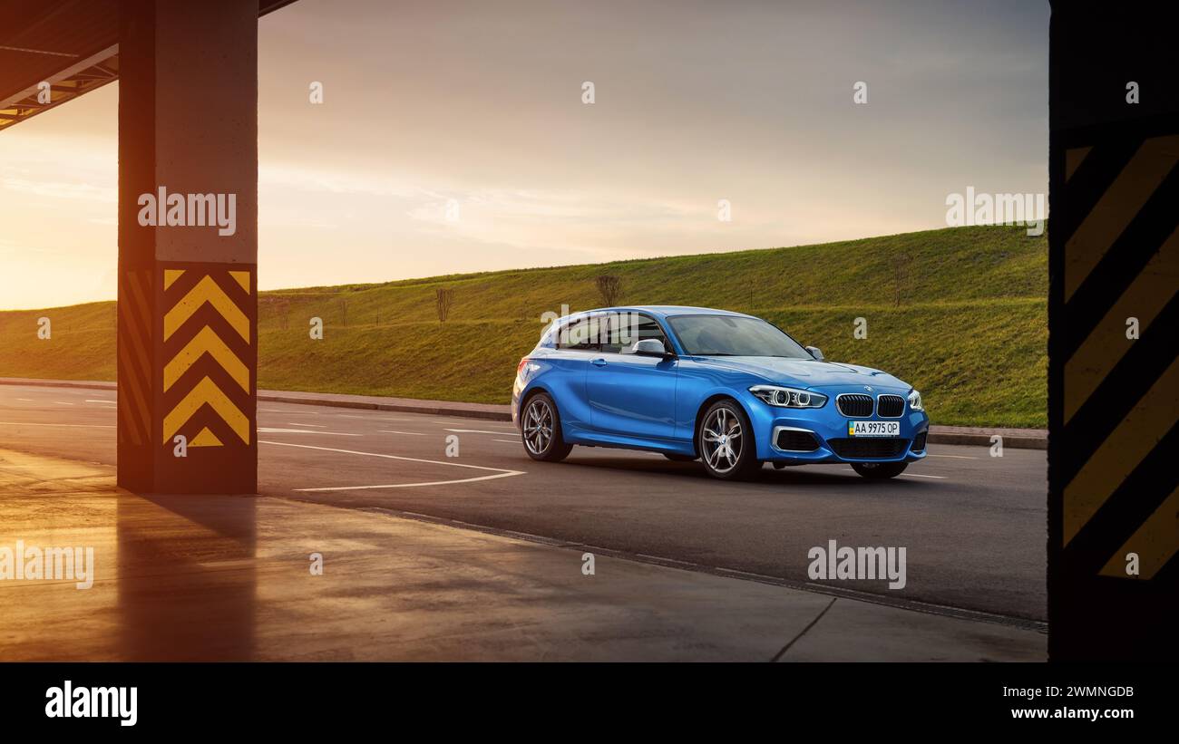 Blauer BMW 1er (F21) auf einem Parkplatz bei Sonnenuntergang. Dreiviertelansicht der sportlichen BMW M140i Fließheck mit zwei Säulen im Vordergrund Stockfoto