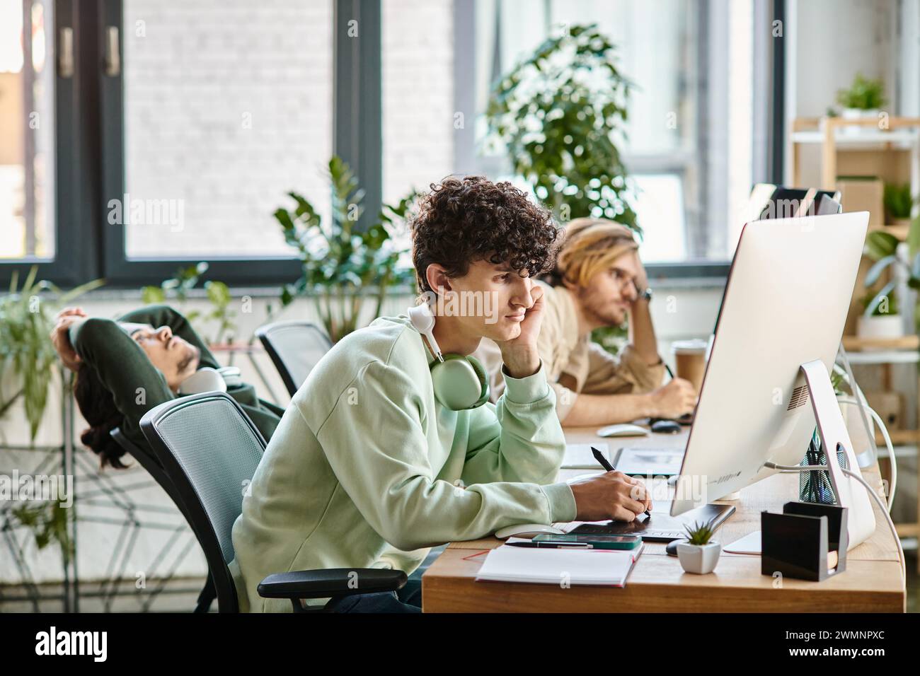 Junger Mann mit lockigen Haaren, der Fotos auf einem Tablet bearbeitet und in einem modernen Startup-Büro auf den Monitor blickt Stockfoto