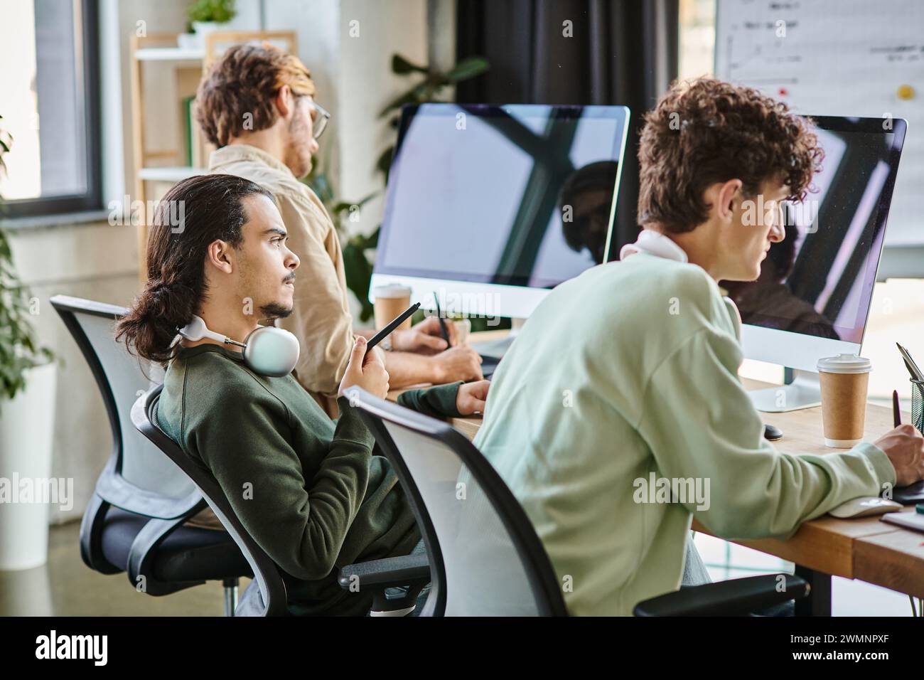 Junger Mann mit Kopfhörern, der Fotos auf dem Tablet bearbeitet und den Monitor im Startup-Büro betrachtet Stockfoto