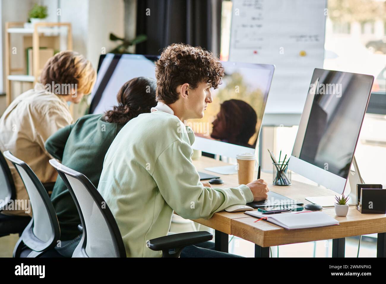 Junger Mann mit lockigen Haaren, der Fotos auf dem Tablet bearbeitet und den Monitor im Startup-Büro betrachtet Stockfoto