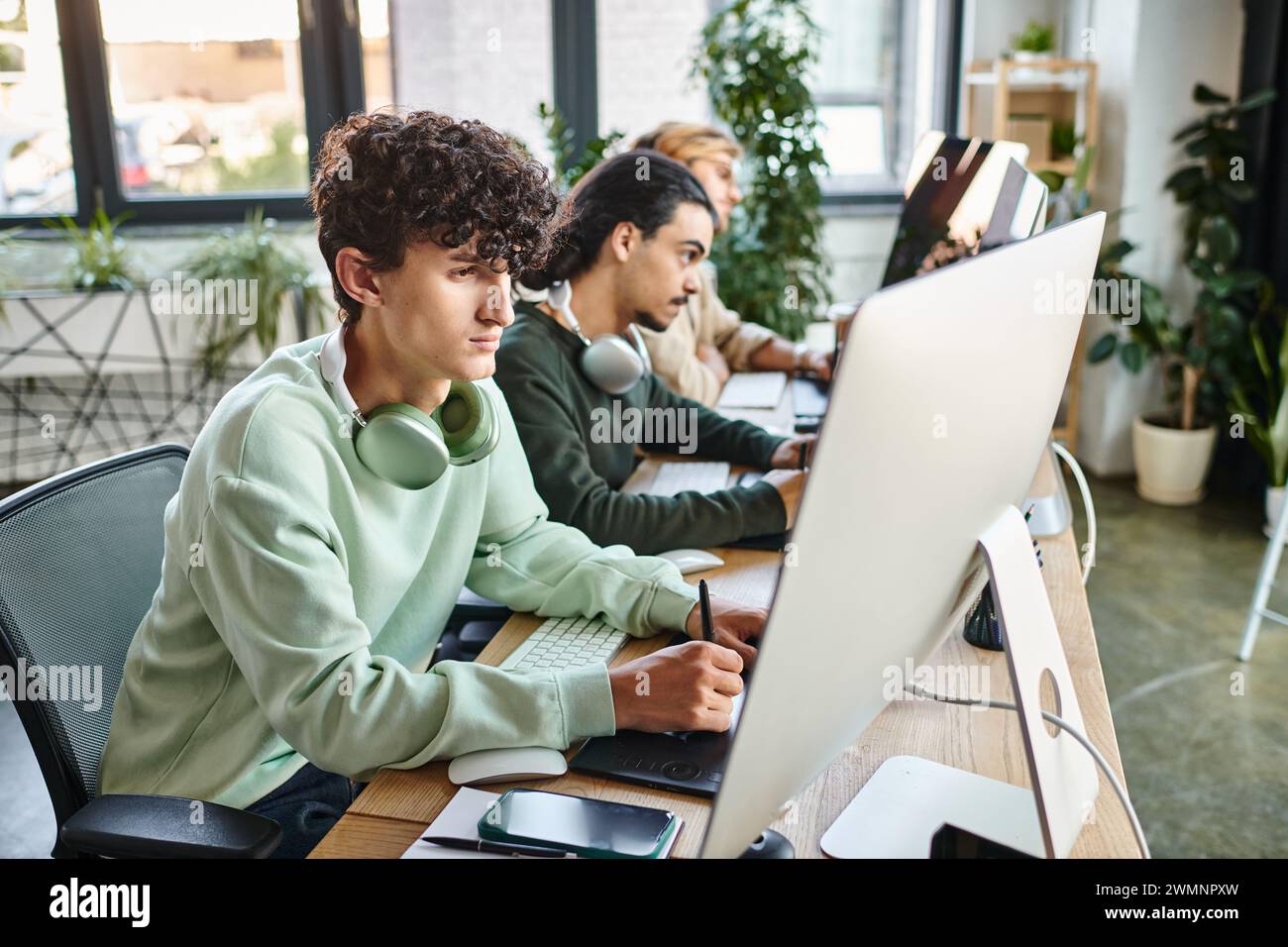 Fokussierter Mann mit lockigen Haaren, der Fotos auf dem Tablet bearbeitet und den Monitor in einem modernen Startup-Büro betrachtet Stockfoto