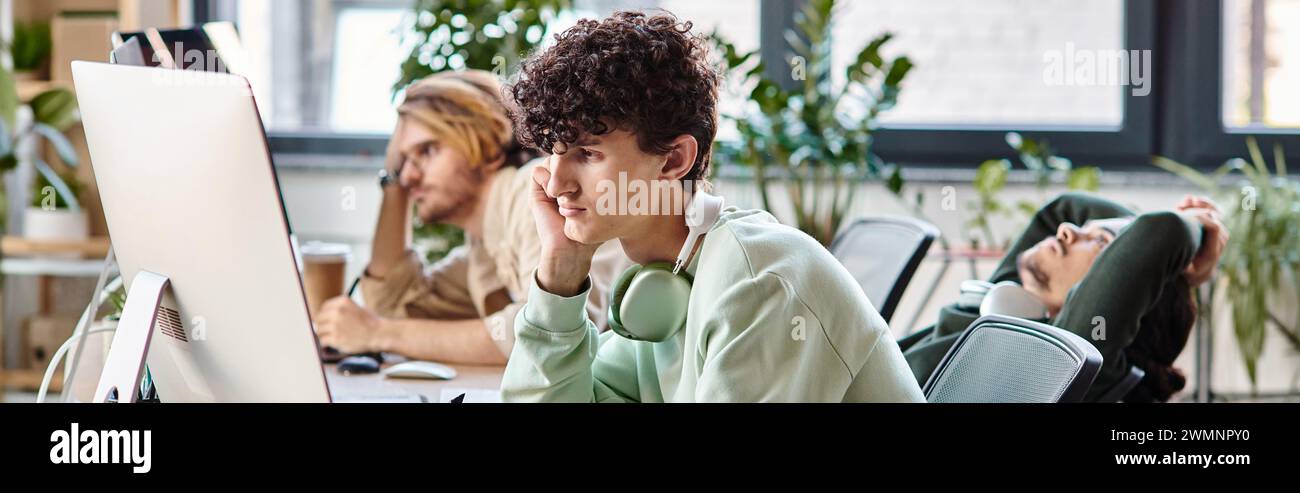 Junger Mann mit lockigen Haaren, der Fotos auf dem Tablet bearbeitet und auf den Monitor im Startup-Büro blickt, Banner Stockfoto