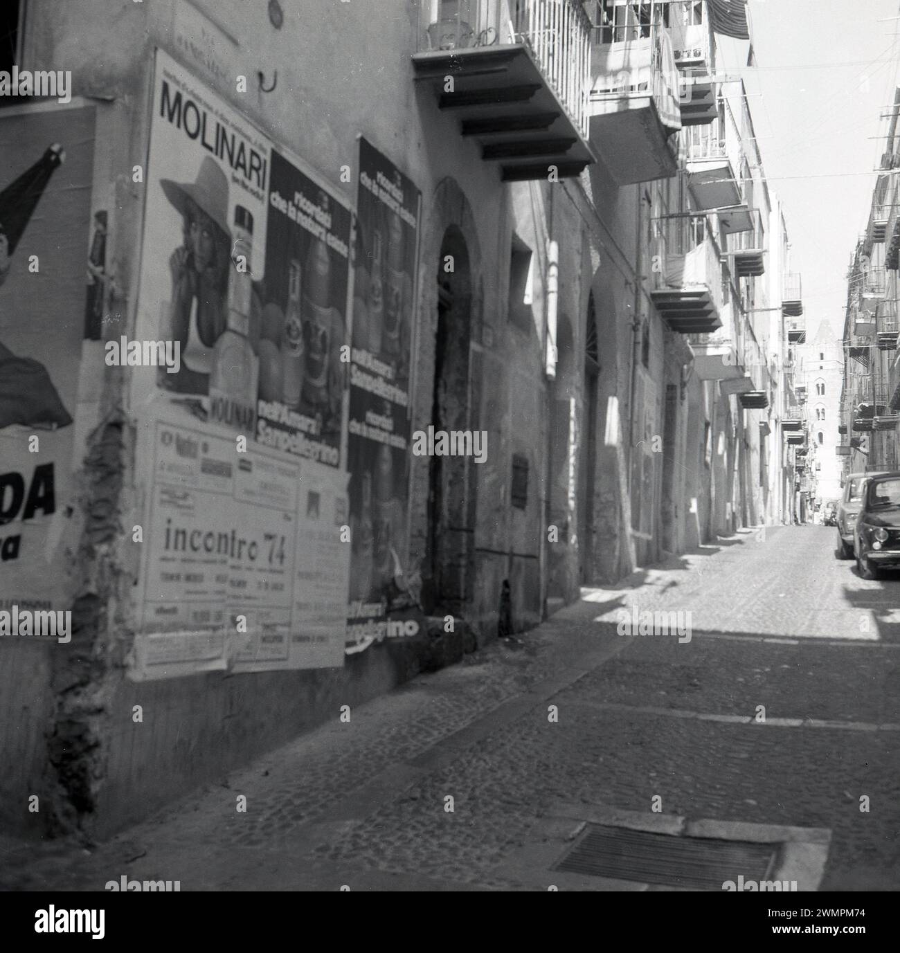 1970er Jahre, historisch, Seitenstraße, Via Mandralisca, Cefalu, Sizilien, Italien... Werfen Sie Plakate an einer Wand für Molinari, einen italienischen Likör, San Pellegrino Water und eines für die Jazzband Inconrtro '74, die besagen, dass Tickets an der Piazza Umberto, dem berühmten Platz im Zentrum von Capri, erhältlich sind. Ein Fiat Punto parkte in der engen Kopfsteinpflasterstraße. Stockfoto