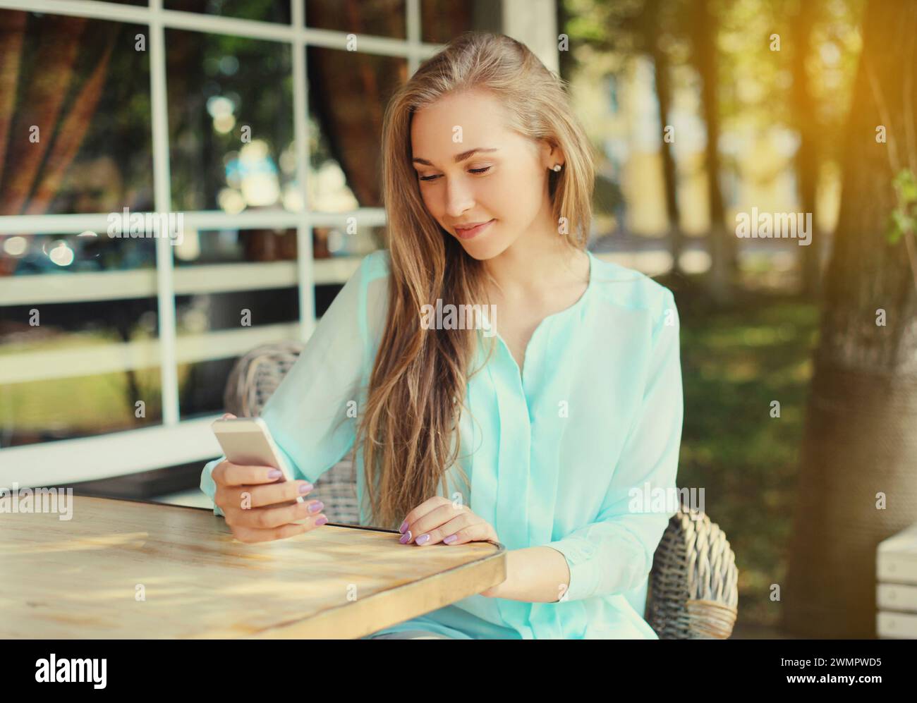 Glückliche kaukasische junge Frau 20s mit Handy, die auf das Gerät schaut, während sie am Tisch im Straßencafé im Sommerpark sitzt Stockfoto