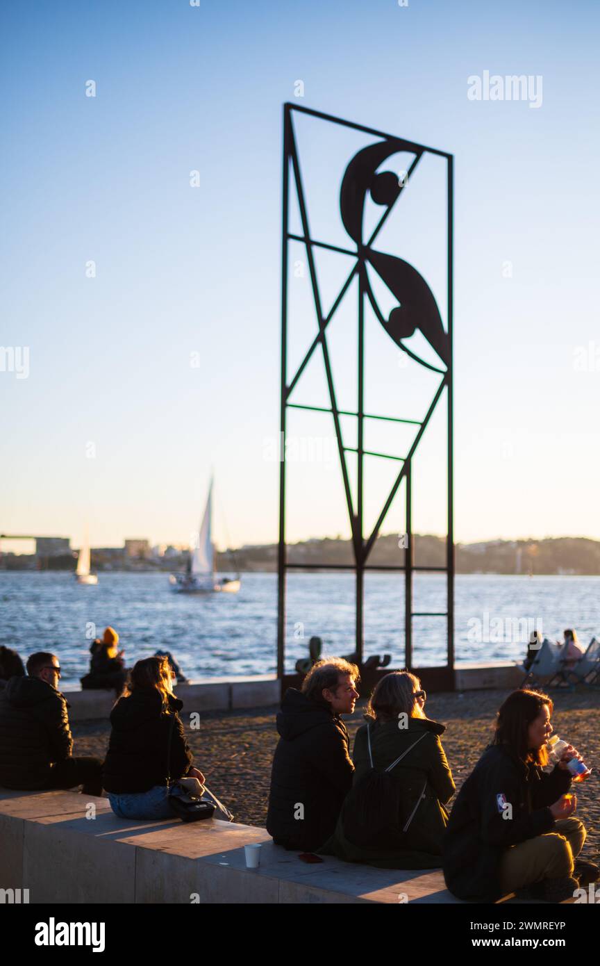 Menschen genießen den Sonnenuntergang auf Ribeira das Naus, die Uferpromenade und den städtischen Strand, und Reminiscência (Almada Negreiros) Skulptur, Lissabon, Hafen Stockfoto