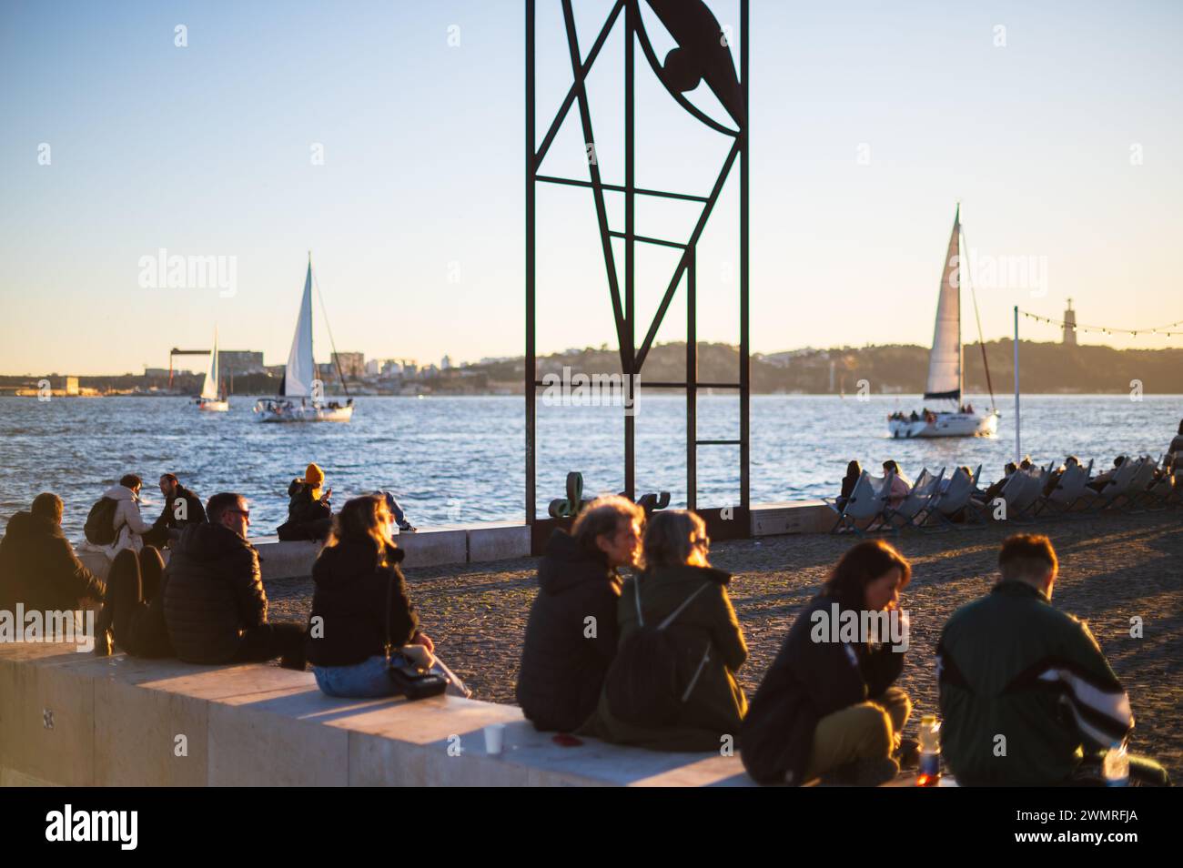 Menschen genießen den Sonnenuntergang auf Ribeira das Naus, die Uferpromenade und den städtischen Strand, und Reminiscência (Almada Negreiros) Skulptur, Lissabon, Hafen Stockfoto