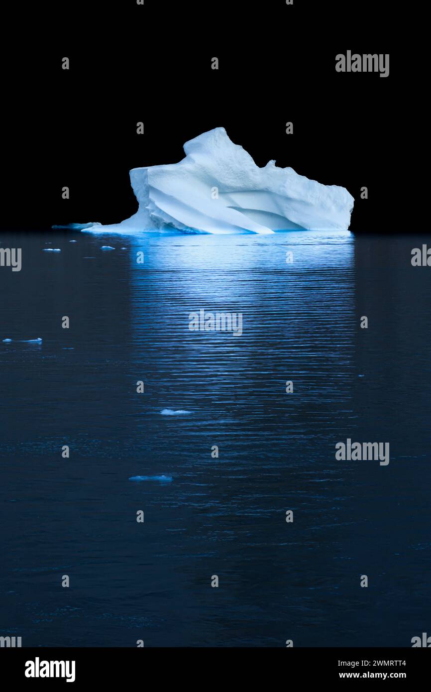 Exquisit geschnitzter Eisberg schwimmt vor dunklem Hintergrund und erzeugt wunderschöne blaue Wellen im Meer. Ostgrönland. Stockfoto