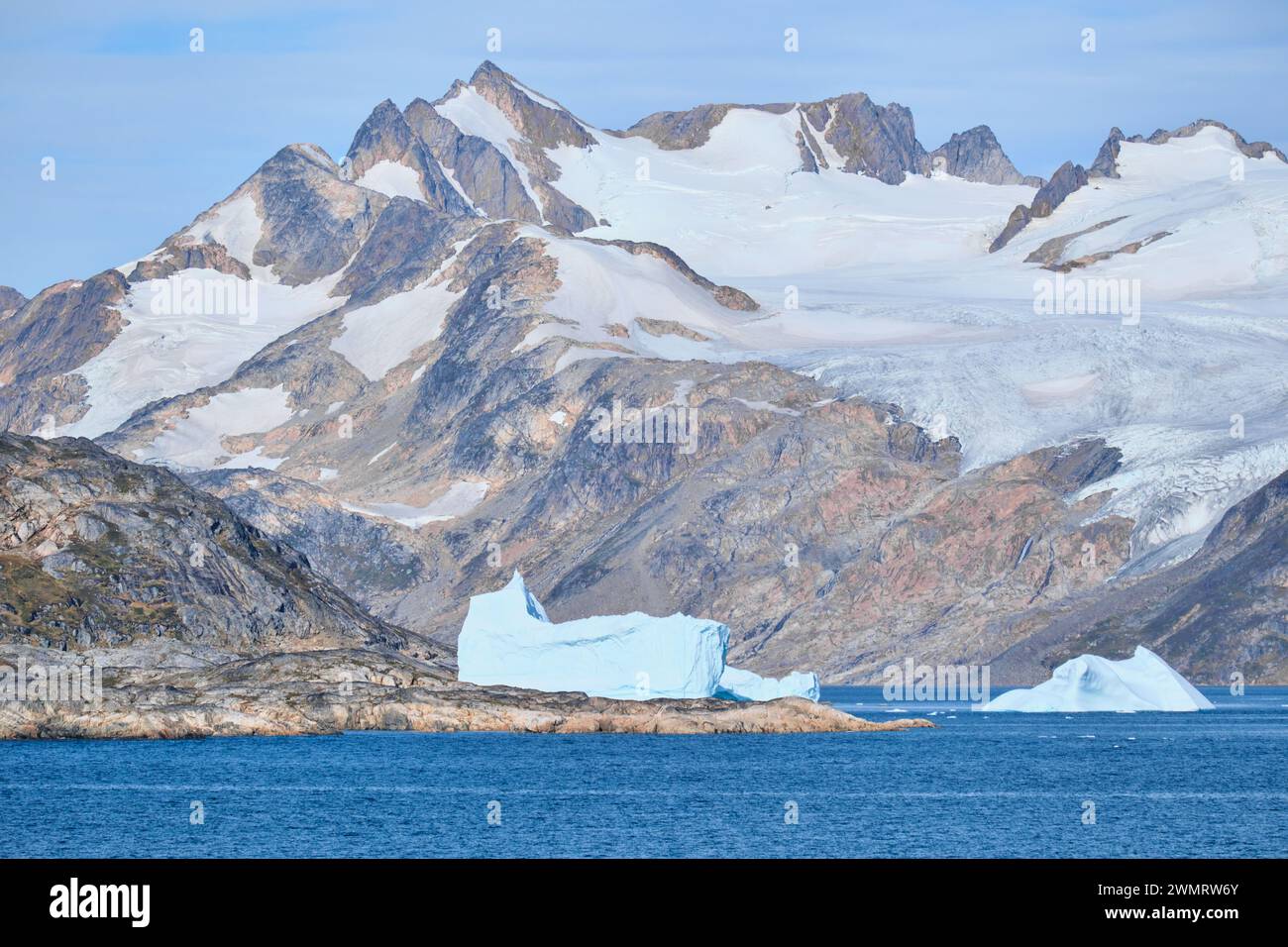 Große Eisberge schweben im strahlend blauen Meer neben einem massiven, zerklüfteten Berg, der mit Schnee und Eis bedeckt ist. Stockfoto