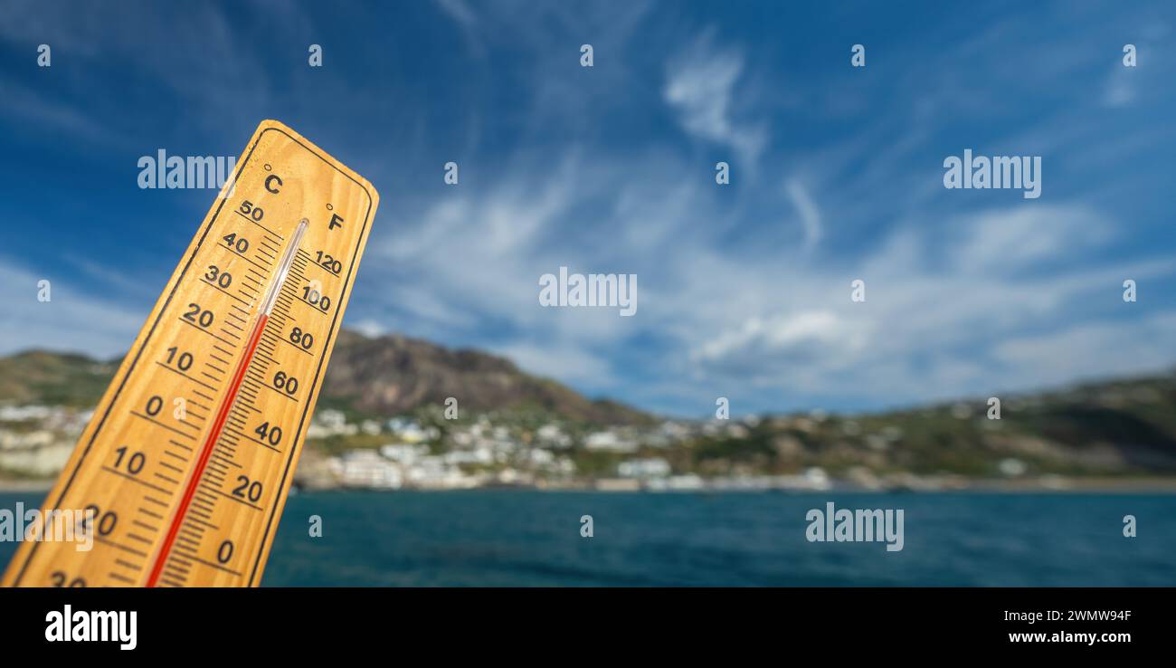 Holzthermometer mit roter Messflüssigkeit, die hohe Temperaturen an sonnigem Tag vor dem Hintergrund des Meeres anzeigt. Konzept von Urlaub, Urlaub, warmes Wetter Stockfoto