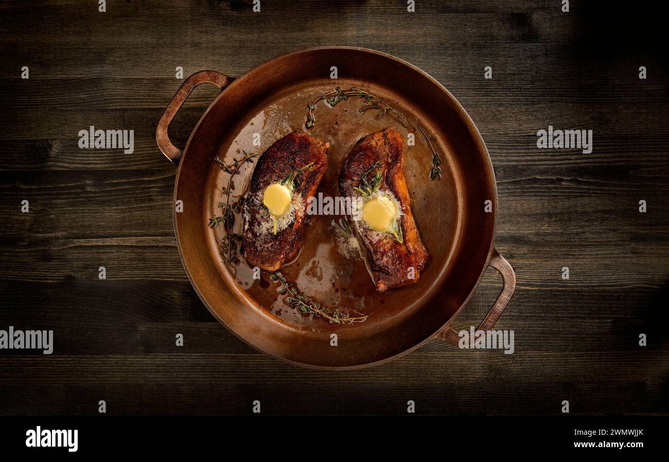 Ein Blick von oben auf zwei appetitliche Striploin-Steaks, die in einer gusseisernen Pfanne mit Butter und Rosmarinkräutern zubereitet werden Stockfoto