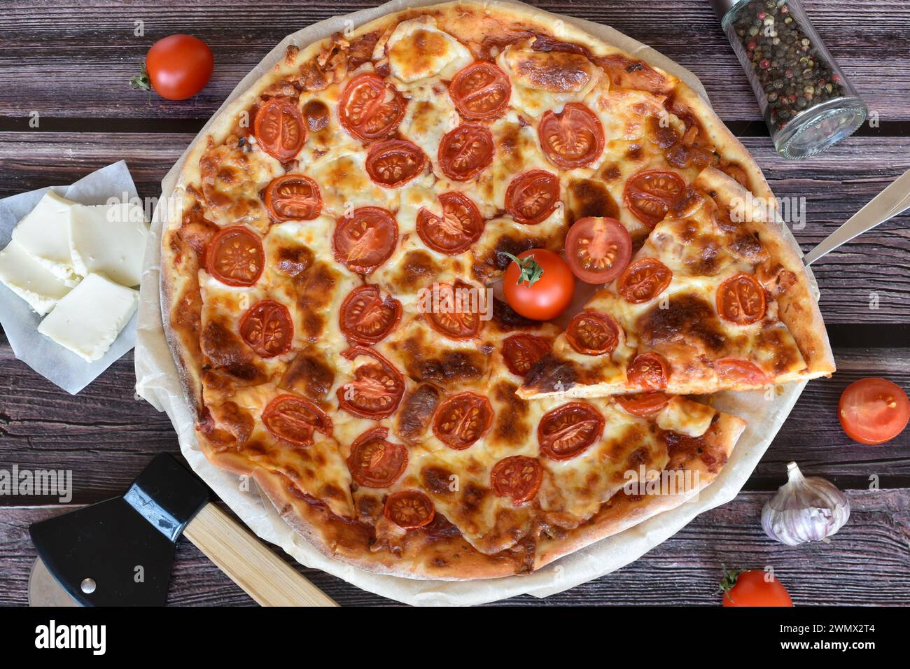 Schaufel mit Pizzascheibe. Pizza Margherita auf rustikalem Holztisch. Kirschtomaten, Mozzarella, Pizzaschneider, Pfeffermühle, Knoblauch. Stockfoto