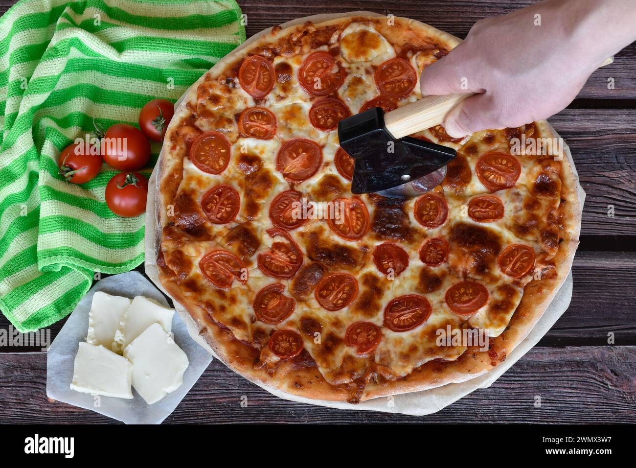 Pizzaschneider in der Hand, frisch gebackene hausgemachte Pizza Margherita serviert auf Holztisch. Rote Kirschtomaten, Mozzarella, gestreifte Serviette. Stockfoto