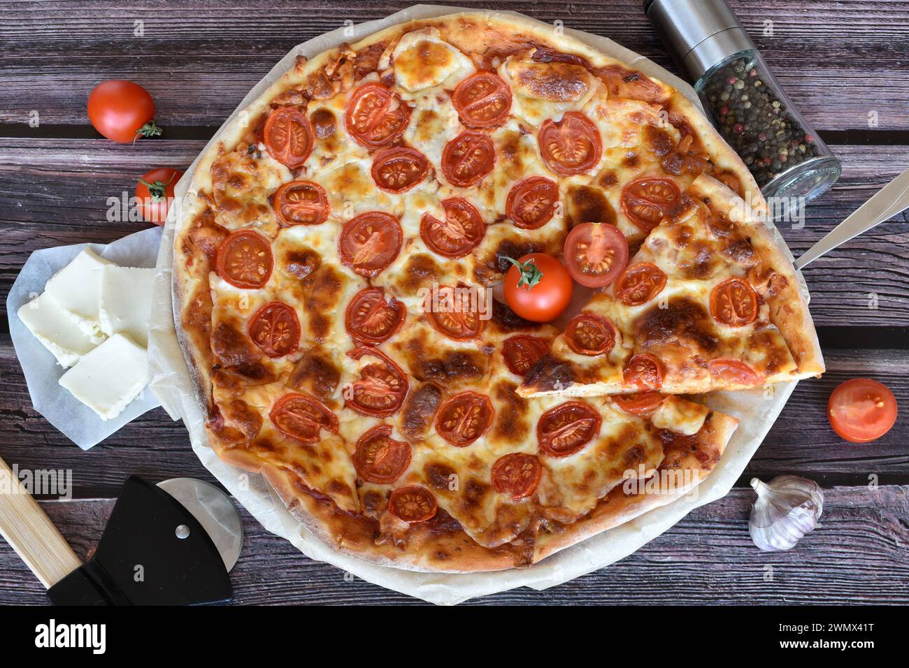 Schaufel mit Pizzascheibe. Pizza Margherita auf rustikalem Holztisch. Kirschtomaten, Mozzarella, Pizzaschneider, Pfeffermühle, Knoblauch. Stockfoto