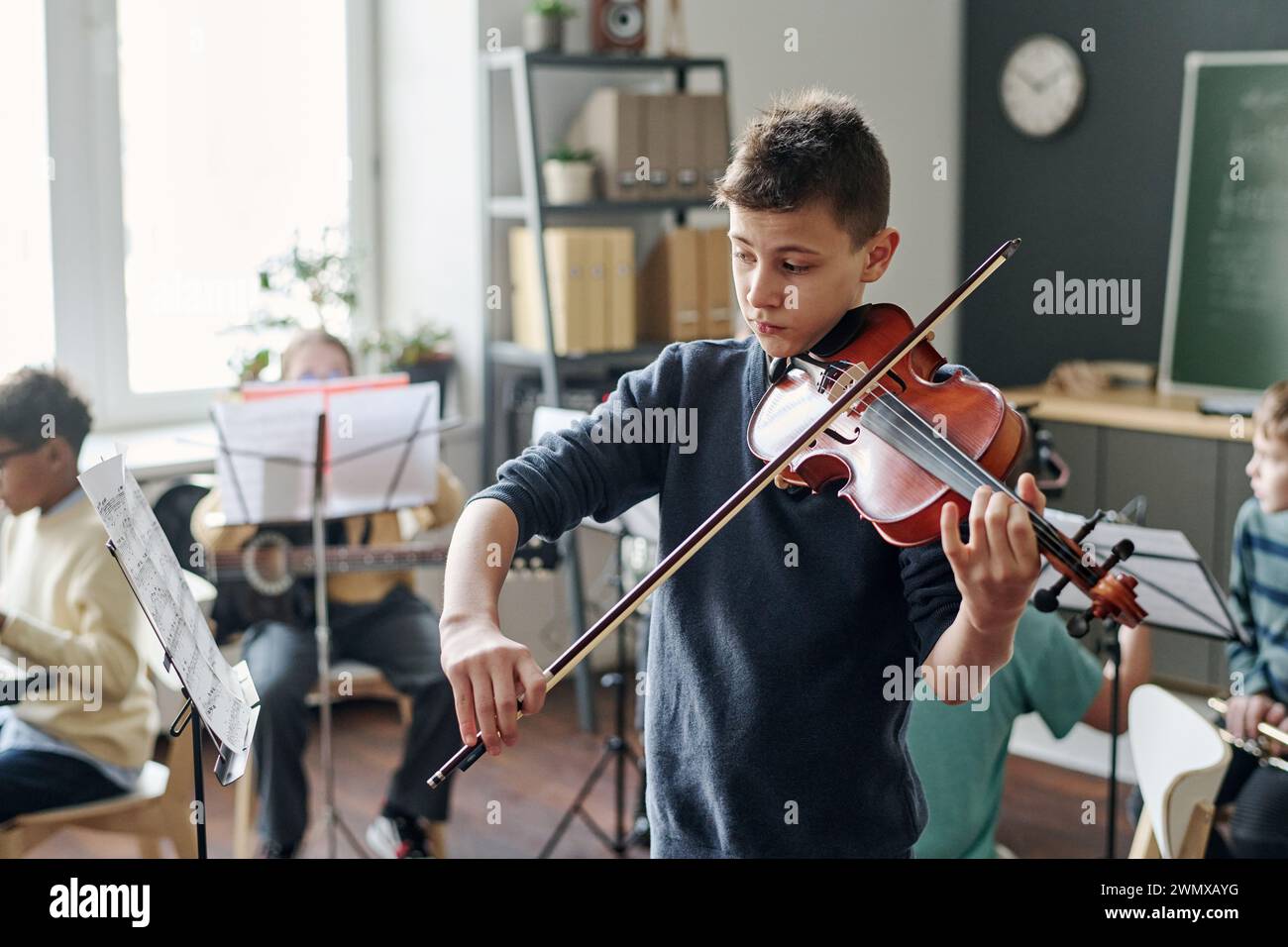 Mittlere Aufnahme eines kaukasischen Jungen, der im Klassenzimmer Geige für Schulorchester übt, seine Klassenkameraden sitzen im Hintergrund Stockfoto