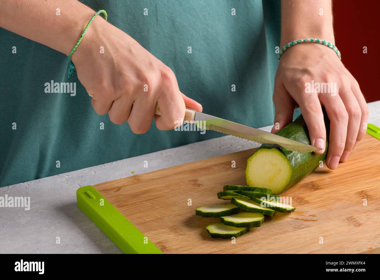 Authentische weibliche Hände schneiden Zucchini auf hölzernem Schneidebrett auf Küchentisch. Frau in Schürze geschnittene Zucchini für die Zubereitung von Lebensmitteln. Vegan und vegetarisch Stockfoto