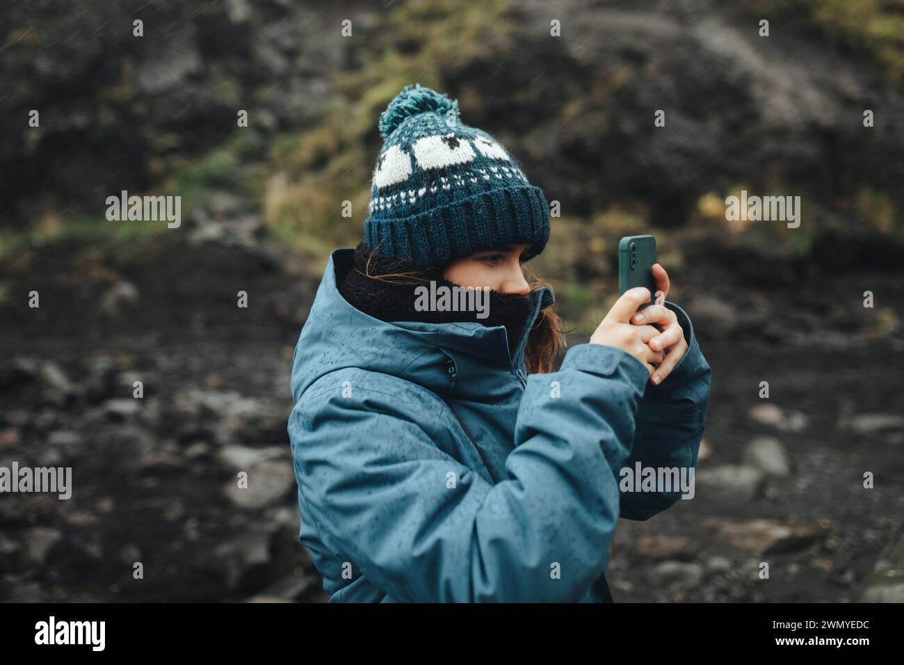 Seitenansicht einer Frau in einem warmen Hut mit einem Smartphone, um isländische Landschaften an einem bewölkten Tag festzuhalten. Stockfoto