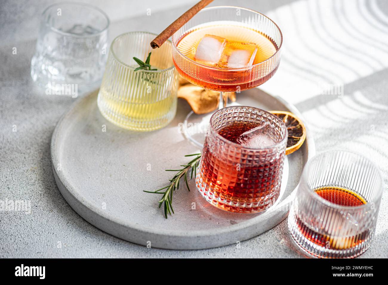 Eine stilvolle Auswahl an alkoholischen Getränken in verschiedenen Gläsern mit Zimtstab, Rosmarin und Eis auf einem runden Tablett aus Beton in weicher Form Stockfoto