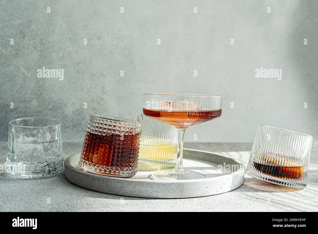 Eine Auswahl an alkoholischen Getränken in strukturierten Gläsern, elegant auf einem runden Betontablett mit sanftem grauen Hintergrund Stockfoto
