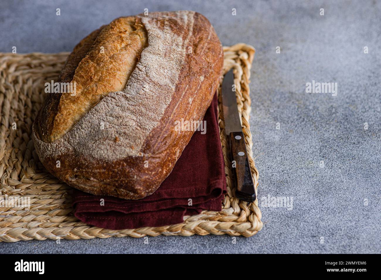 Ein knuspriger Brotlaib mit selbstgemachtem Roggensauerteig, auf einer gewebten Matte mit einer dunkelroten Serviette und einem Brotmesser Stockfoto