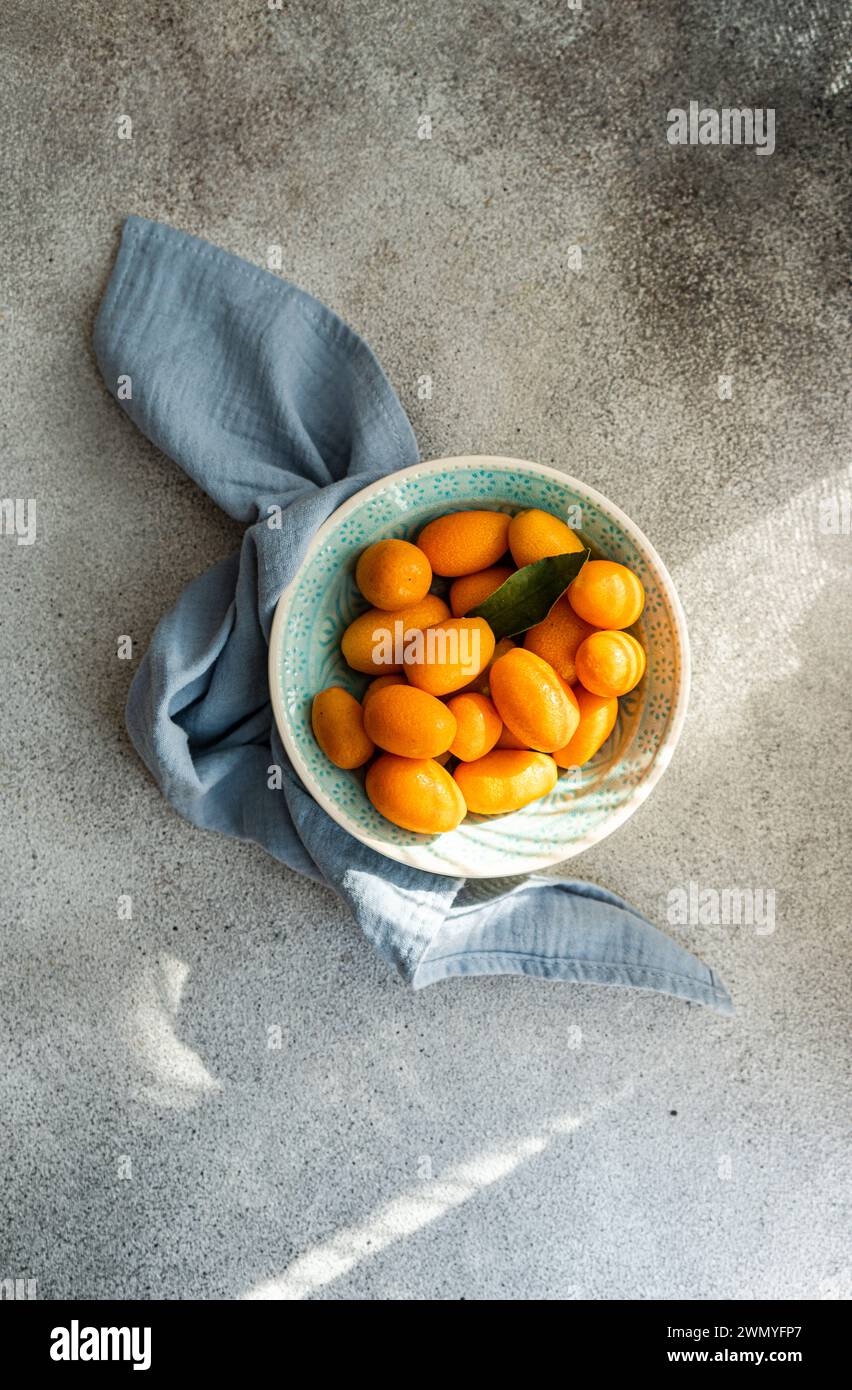Von oben frische Kumquats in einer dekorativen Schüssel mit einer blauen Serviette auf einem strukturierten Betonhintergrund Stockfoto