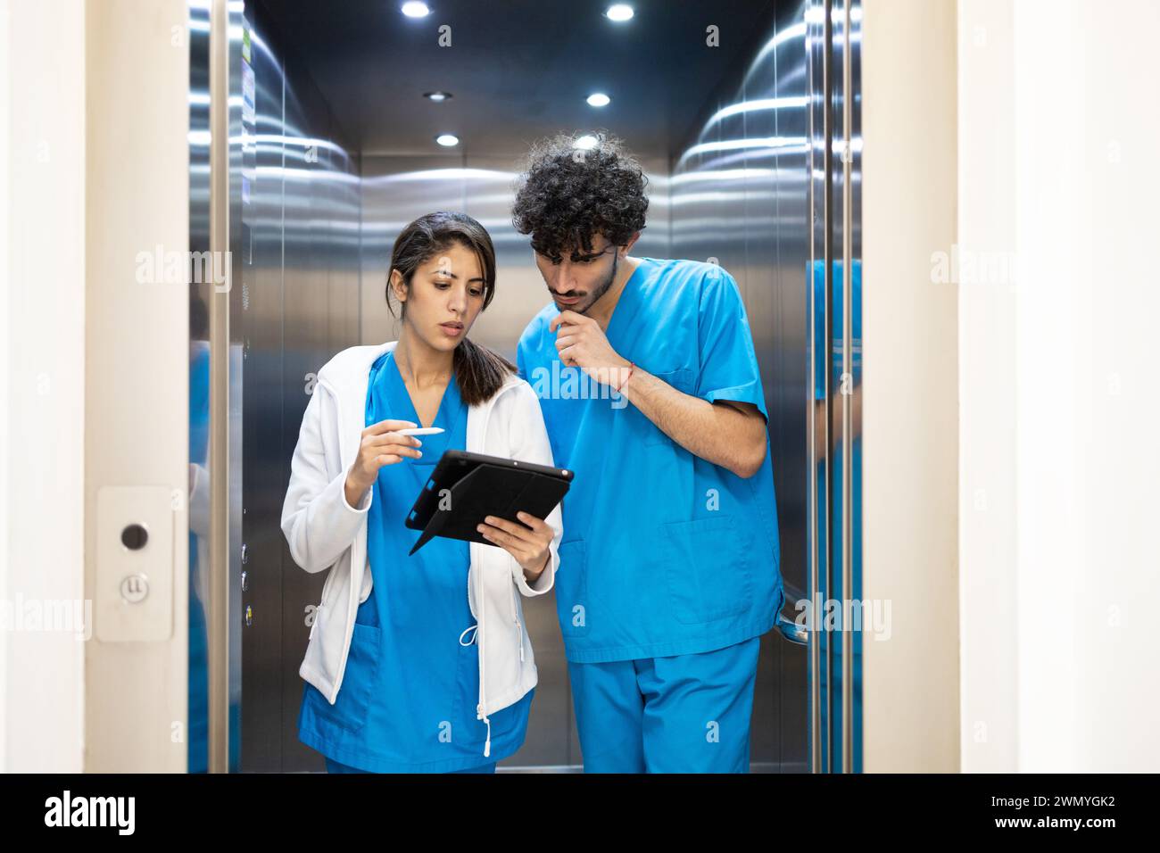 Zwei medizinische Fachkräfte in blauen Peelings überprüfen medizinische Aufzeichnungen auf einem Tablet in einem gut beleuchteten Krankenhausaufzug. Stockfoto