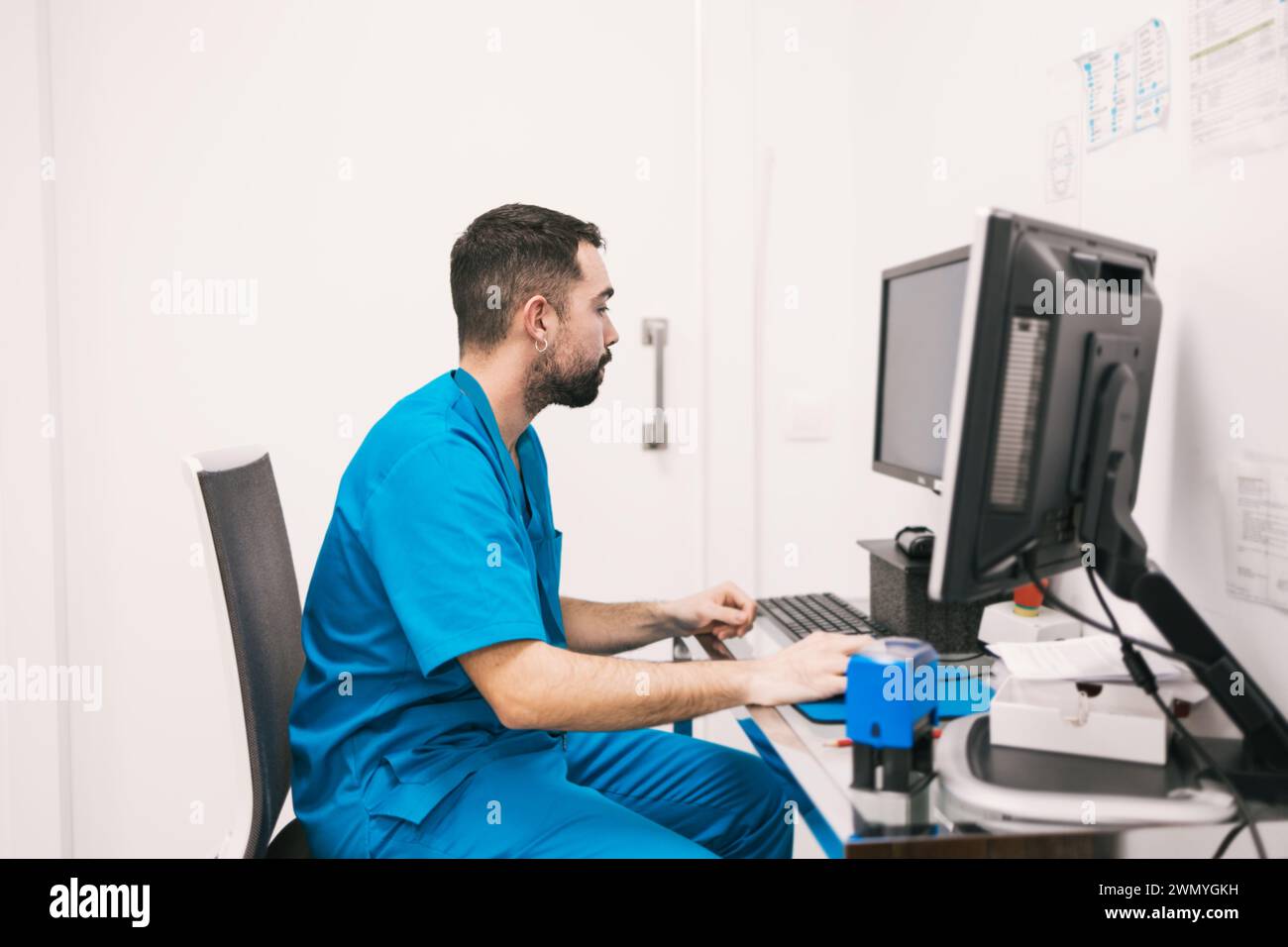 Eine engagierte männliche Krankenschwester in Peelings konzentriert sich auf die Aktualisierung von Patientenakten auf einem Computer in einer medizinischen Klinik. Stockfoto