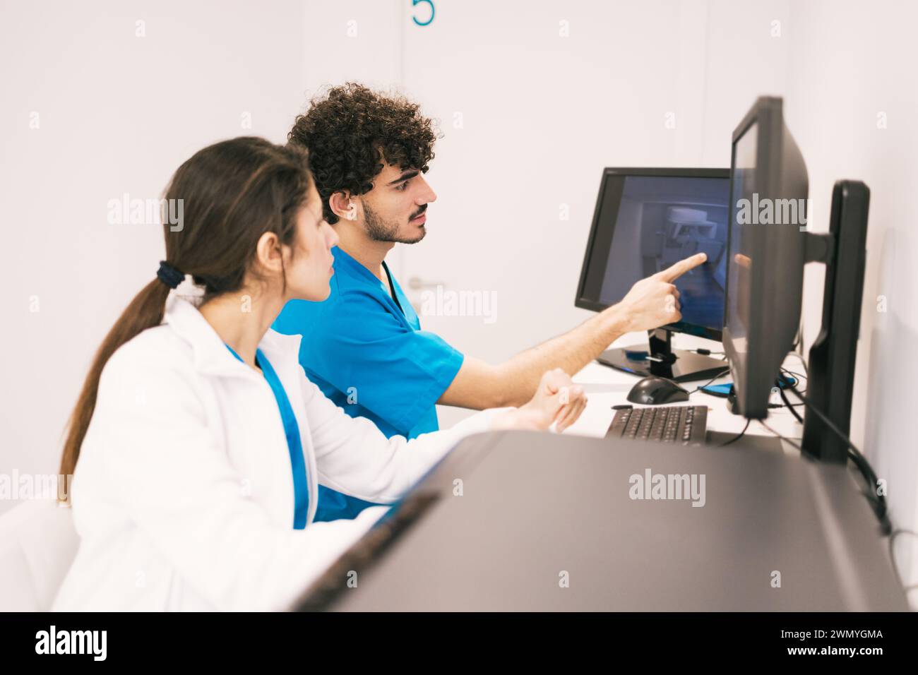 Zwei medizinische Fachkräfte in blauen Peelings konzentrieren sich auf einen Computerbildschirm und diskutieren medizinische Daten in einem hellen Büro. Stockfoto
