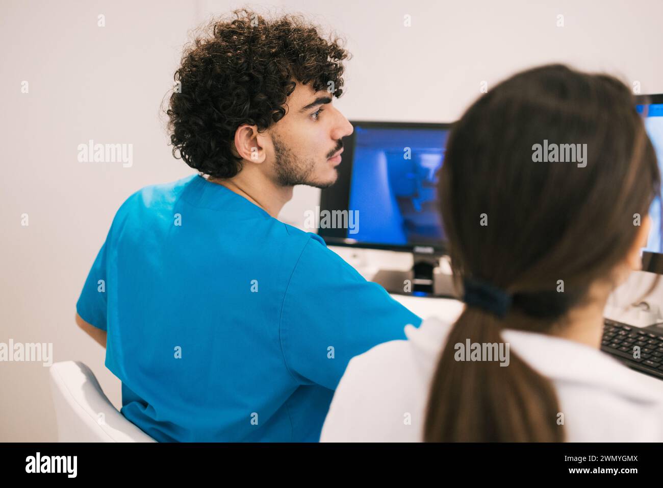 Ein männlicher medizinischer Mitarbeiter in blauem Scrubs bespricht Patientendaten mit einer Kollegin vor einem Computerbildschirm in einer klinischen Umgebung Stockfoto
