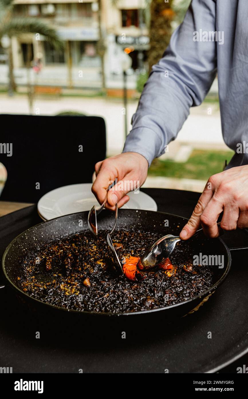 Eine Person serviert eine Portion traditioneller spanischer schwarzer Paella, die in einer großen, flachen Pfanne gekocht wird und die appetitlichen Meeresfrüchte hervorhebt Stockfoto