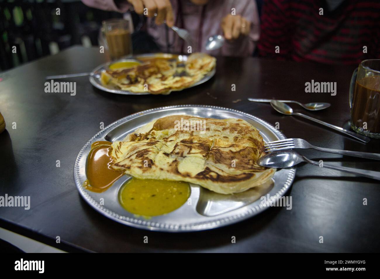 Malaysische Küche Grundnahrungsmittel, Roti Canai, serviert auf Metalltellern mit Dhal und Curry, während die Leute Tee im Hintergrund genießen. Stockfoto