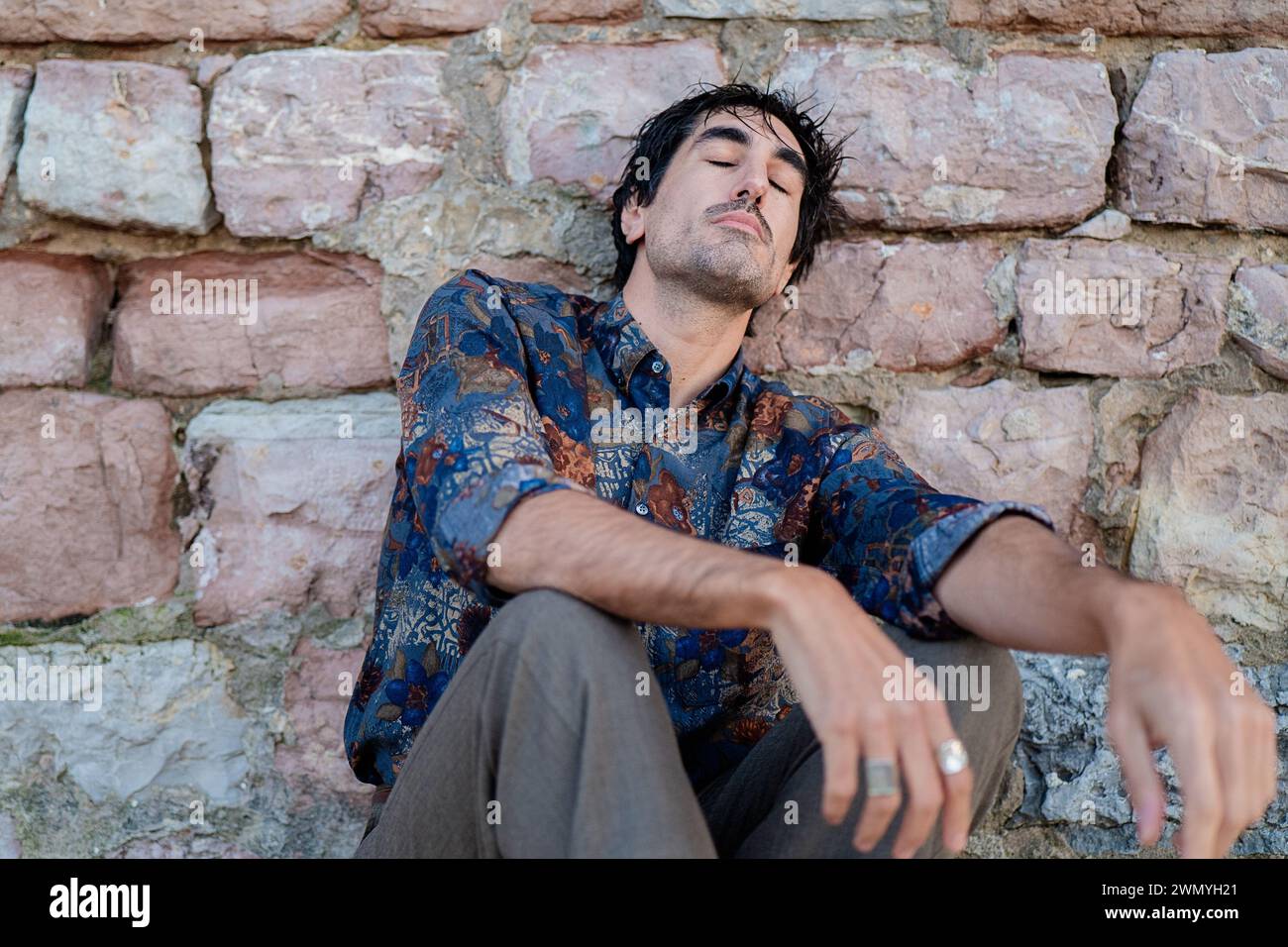 Ein junger Mann mit nachdenklichem Ausdruck sitzt an einer alten Steinmauer und erscheint verloren in Gedanken Stockfoto