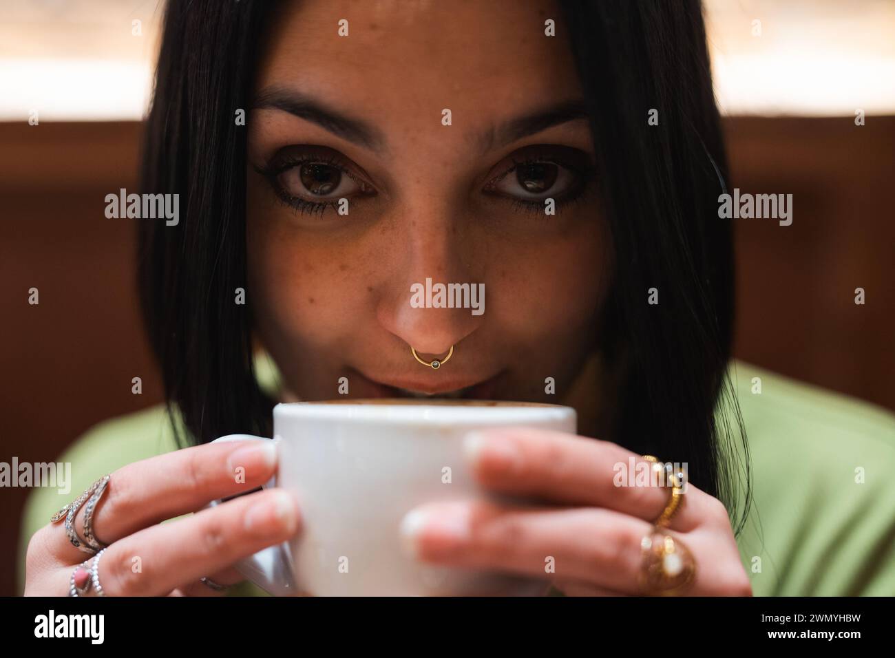 Eine Nahaufnahme einer Frau, die eine warme Tasse Kaffee genießt, geschmückt mit zartem Schmuck und einem Nasenring, während sie nachdenklich schaut. Stockfoto