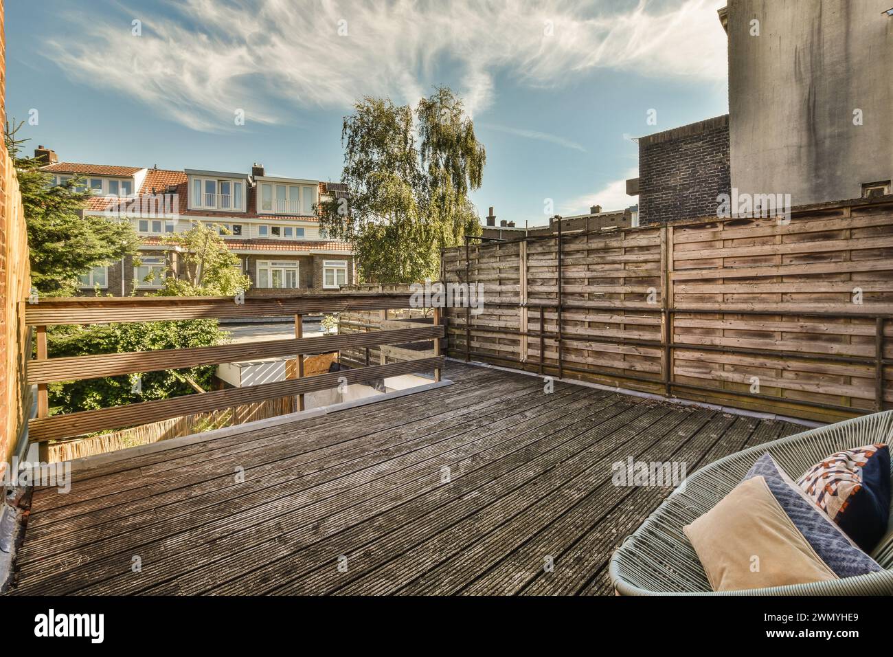 Eine einladende, urbane Dachterrasse mit Holzterrasse, komfortablen Sitzgelegenheiten und üppigen grünen Bäumen vor dem Hintergrund von Vorstadthäusern unter einem blu-ray Stockfoto