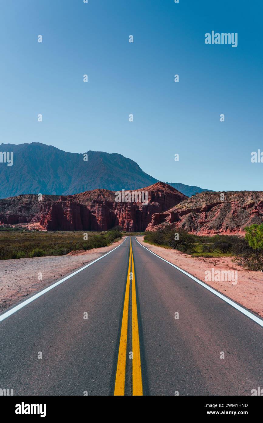 Gerade Straße, die sich in Richtung roter Felsformationen unter einem riesigen blauen Himmel in Los Castillos im Norden Argentiniens erstreckt Stockfoto