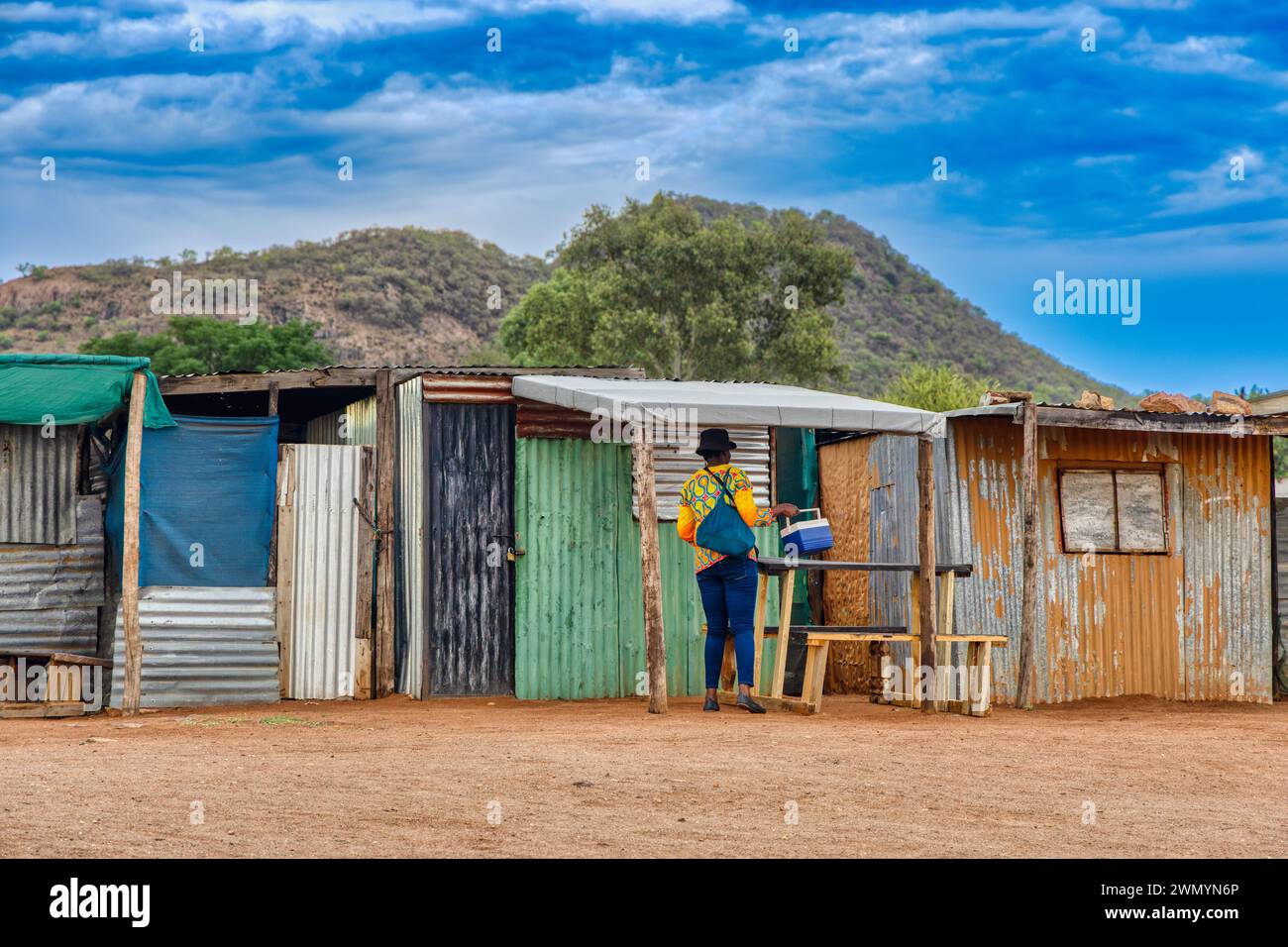 township informelle Siedlung in afrika in der Nähe eines Hügels, Frau, die eine Kühlbox vor den Wellblechhütten pflegt Stockfoto