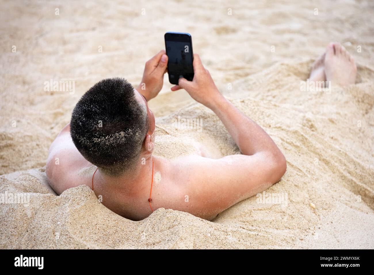 Mann, der in einem Sand begraben liegt, mit Smartphone in den Händen. Urlaub und Online-Kommunikation am Strand Stockfoto