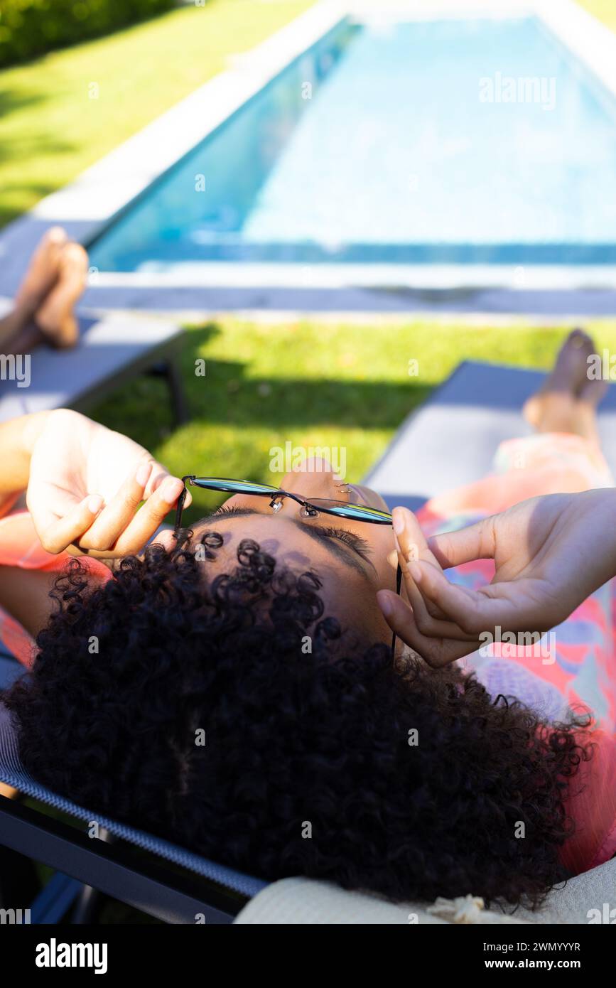 Eine junge Frau, die sich auf einer Sonnenliege am Pool entspannt, mit Sonnenbrille in der Hand, mit Kopierraum Stockfoto
