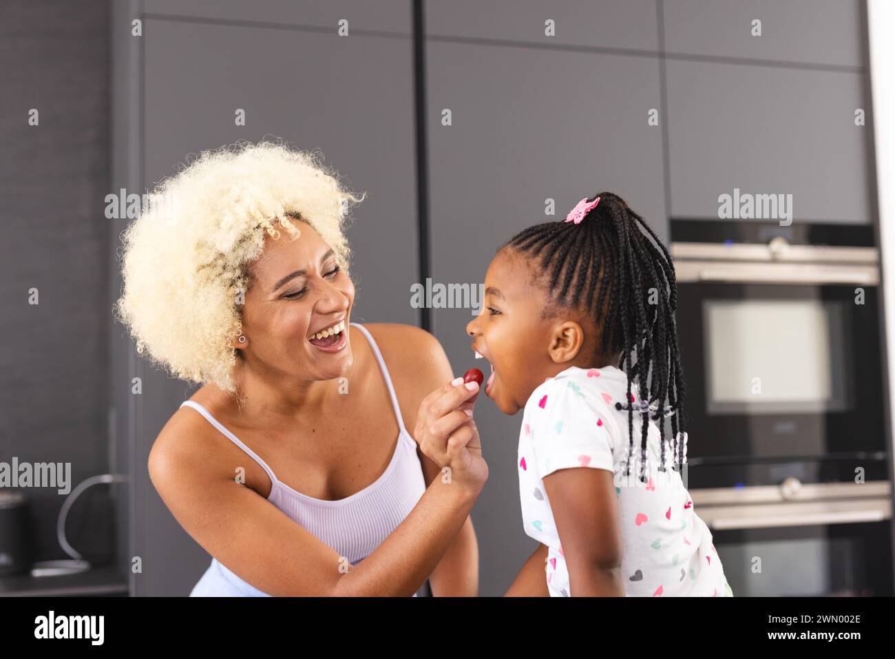 Junge birassische Mutter mit blonden lockigen Haaren lacht mit einer afroamerikanischen Tochter in einer Küche Stockfoto