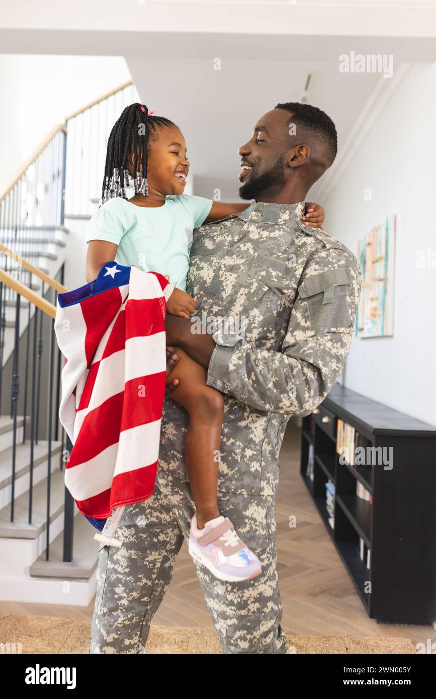 Ein Soldat in Uniform hält ein junges Mädchen zu Hause und drückt Freude mit einer amerikanischen Flagge aus Stockfoto