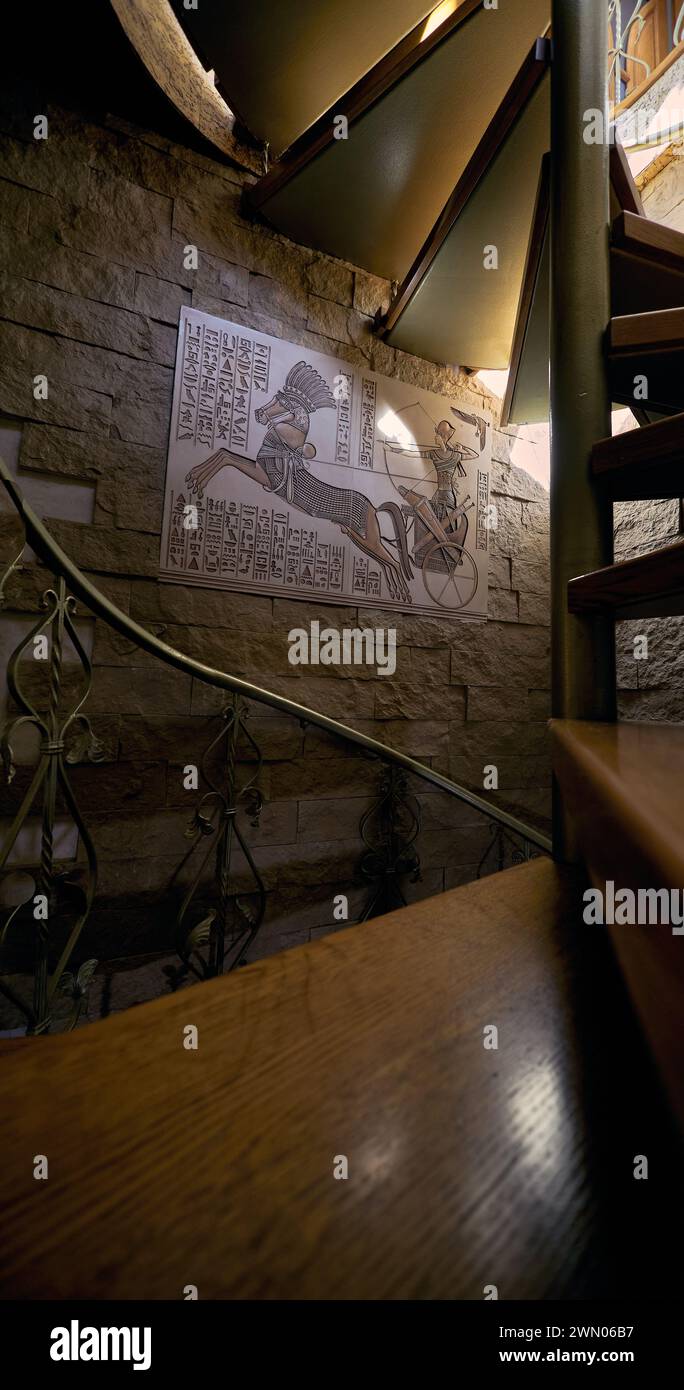 Inneneinrichtung. Pano mit einem ägyptischen Pharao auf einem Wagen an der Wand in der Nähe einer spiralförmigen Metalltreppe mit Holztreppen und Metalldekoration Stockfoto