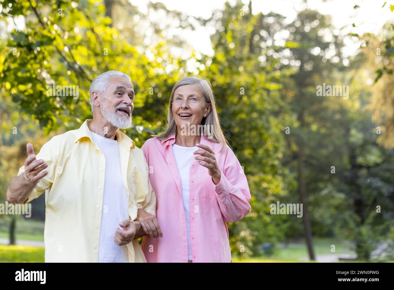 Fröhliches älteres Ehepaar, das sich bei einem gemütlichen Spaziergang in einem grünen Park in goldenem Sonnenlicht in einem herzzerreißenden Gespräch unterhält. Stockfoto