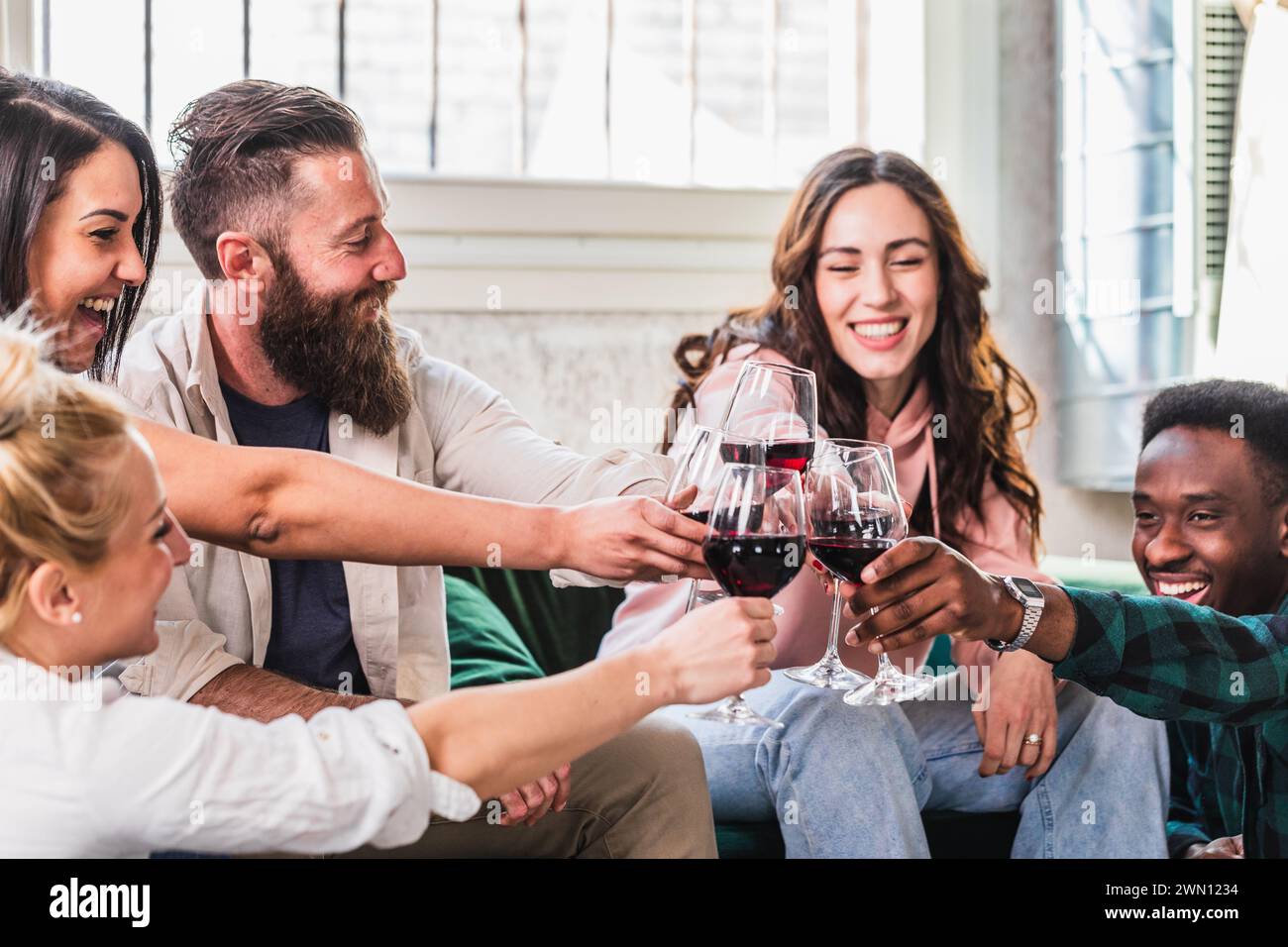 Eine lebhafte Gruppe von Freunden tobt mit Rotwein und teilt einen Moment der Freude und der Feier in einem hell beleuchteten Wohnzimmer, um das Gefühl des Kameraden zu fördern Stockfoto