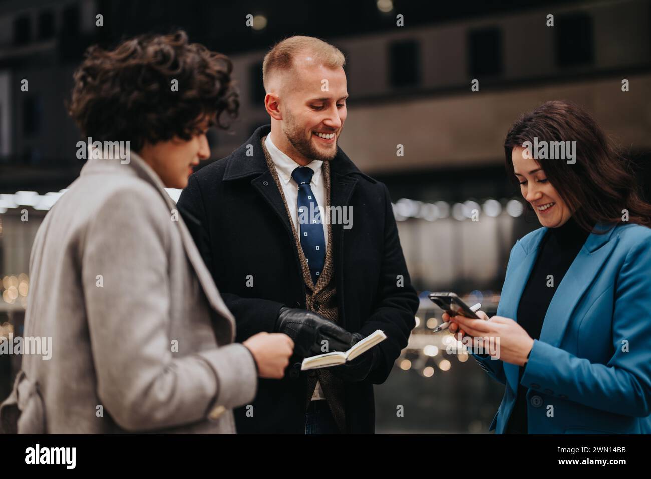 Drei Personen in stilvoller Winterkleidung teilen sich einen Moment der Freude, wenn sie ein Smartphone auf einer Straße der Stadt betrachten, das Teamwork und Konnektivität ausstrahlt. Stockfoto