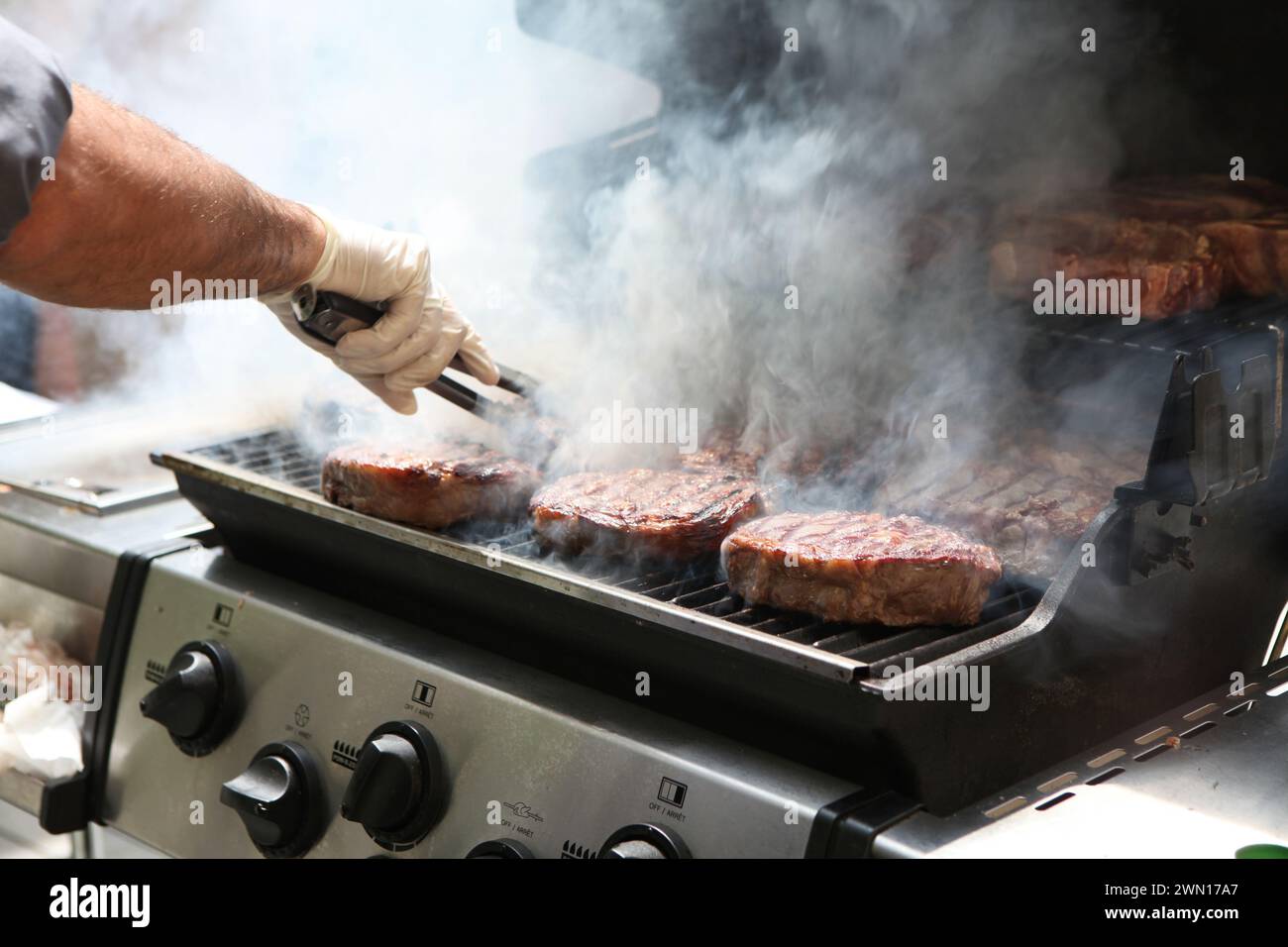 Nahaufnahme einer Hand eines Mannes, der eine Zange hält und Rindersteaks auf einem rauchigen Grill dreht Stockfoto