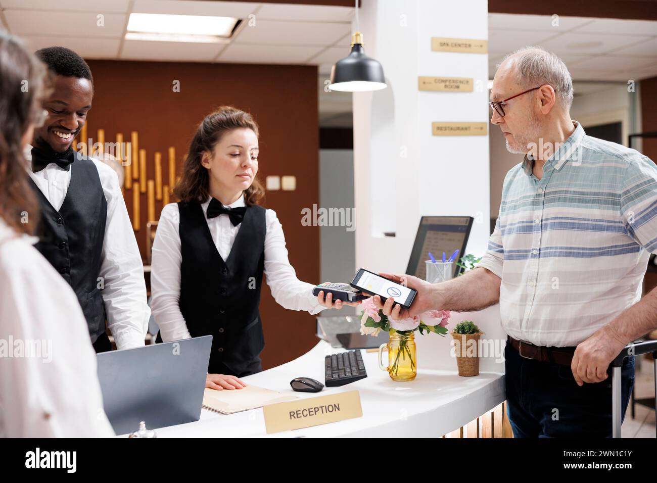 Ein älteres Paar meldet sich an der Hotelrezeption an, unterstützt durch freundliches, multiethnisches Personal. Kaukasischer alter Mann an der Rezeption bezahlt am POS Terminal mit Smartphone für Reservierung und Buchung. Stockfoto