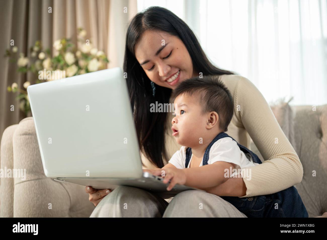 Eine glückliche asiatische Geschäftsfrau, die Mutter, arbeitet von zu Hause aus, arbeitet an ihrem Laptop und kümmert sich um ihren frechen Jungen. Stockfoto